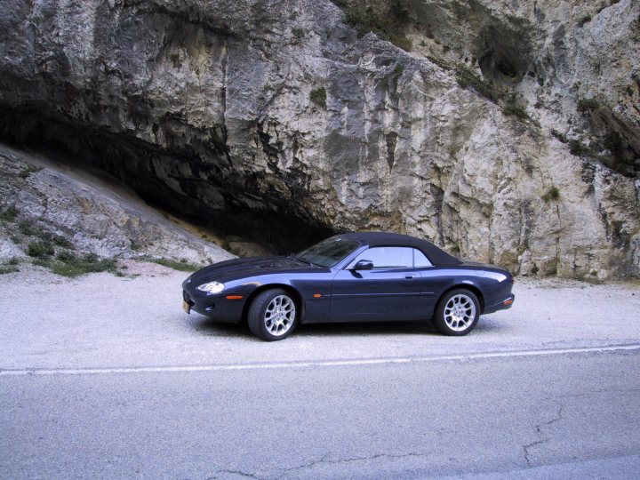 Call Jaguar Roll Pistonheads - The image depicts a dark blue sports car parked on a street next to a natural rock formation. The car, which has a sleek and sporty design, is facing towards the left side of the image. The driver's side door is open, and the car has a convertible top that is not visible in this image. The road curves gently in the background, suggesting that the photographer is on an alpine highway. The rock formation close by adds a dramatic backdrop to the scene.