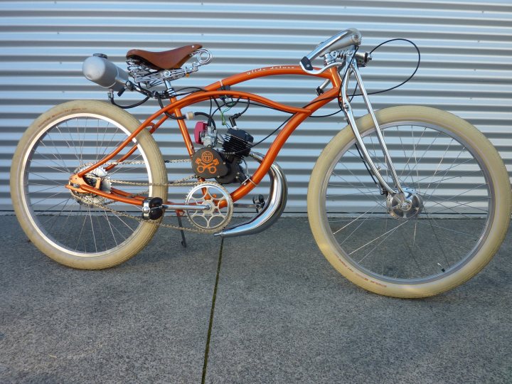 Motorised Bikes - Page 1 - New Zealand - PistonHeads - The image shows a distinctive, rounded, single-wheel trikele against a blue backdrop. The trikele's light orange frame contrasts with its front wheel, which has a gold band and is not visible within the image. On the bike, there's a prominent bobber, which gives the bike its low-riding, high-handling style.

The bike has a unique, avant-garde design, featuring an elongated seat and handlebars. The handlebars are equipped with brakes, suggesting a disc braking system. The trikele's front wheel is covered with a fender, and the seat has a brown leather seat, adding an element of luxury or sportiness.

The close-up view of the trikele from the front showcases its clean design, showcasing the fusion of modern technology with a classic aesthetic. The blue backdrop accentuates the prominence and detail of the bike, making the trikele the focal point of the image.