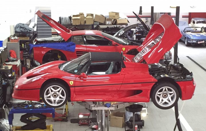 Nearly ready to play........:) - Page 1 - Ferrari V12 - PistonHeads - The image shows a bustling garage bustling with activity and filled with various automotive parts and tools. The main focus is a shiny red Ferrari sports car that's up on a mechanic's lift, its hood opened to reveal its engine. Surrounding the car, there are other vehicles in similar states of disassembly, suggesting ongoing maintenance or repairs. This scene encapsulates the precision and complexity involved in automotive mechanics and restoration.