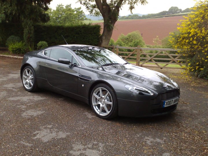 The Proper Colour for an Aston thread - silver, greys, black - Page 1 - Aston Martin - PistonHeads - The image features a sleek, modern car parked on a gravel driveway. It's a blue sports car judging by the sheen and aerodynamic design. The vehicle is positioned facing the camera, with the headlights and grille more visible than the rear of the car. The setting includes a beautiful green or blue lawn in the background, possibly suggesting a rural or countryside location, and the driveway appears to be a private or residential area.