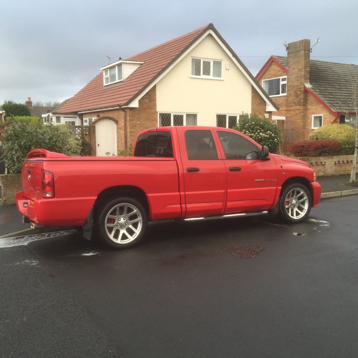 A red truck parked in front of a building - Pistonheads - In the image, there's a red pickup truck parked on a residential street. The truck is facing away from the camera, showcasing the back view. It's situated in front of a white house, which appears to be a two-story building with brick elements. The colors of the truck and house stand out against the grey sky, suggesting that it might be an overcast day. The overall scene gives a quiet, everyday suburban vibe.