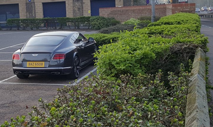 A bit council (Vol 3) - Page 212 - The Lounge - PistonHeads - The image shows a black luxury car parked in a parking space in front of a building. The car is sleek with a clean design and has a license plate. The parking space is marked with white lines, and the building's facade has a mix of brick and stone with closed garage doors. A well-maintained hedge is visible alongside the street where the car is parked.