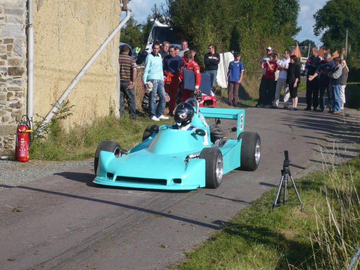 Looking for Radical car - Page 1 - Radical - PistonHeads - The image captures an intense moment at a karting event. A person, wearing a black helmet and a blue suit, is sitting inside a blue and white kart. The kart is on a track, ready to start. A crowd of spectators is standing on the side, eagerly watching the upcoming race. A red fire extinguisher is visible on the ground near the track. Behind the track, there's a brick wall and lush green trees, adding a natural backdrop to the sporting event.