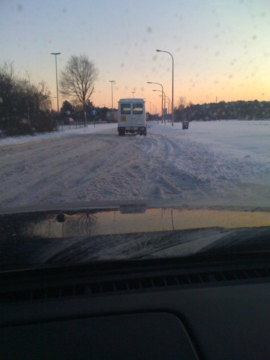 Pistonheads - This image captures a scene on a snowy day. A white vehicle, identified as a delivery truck, is the main subject. The truck is moving down a road, and we see the back end of it, indicating that it is headed towards us. The road itself is covered with a layer of thick snow. The sky above is overcast, suggesting more precipitation to come. Flanking the road are bare trees, their branches devoid of leaves, which are typical for winter. The setting is a quiet street, following the hiatus caused by the weather.