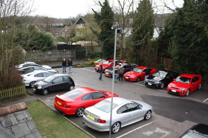 Pistonheads - The image is an aerial or overhead view of a parking lot. The lot is occupied by various cars, including what appears to be sports cars, sports utility vehicles (SUVs), and smaller compacts. People are standing around the area, admiring the cars. There are also some containers and a small white building in the background. The whole scene is framed by external elements such as trees and buildings, indicating that the parking lot is located in an urban or suburban area. The scene suggests that this could be a car show or a gathering of car enthusiasts.