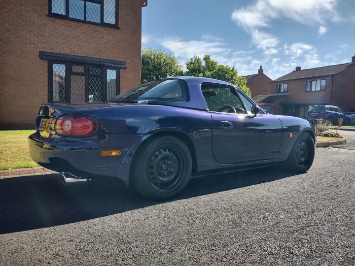 Mx5 1.8 Sport. Had to see what all the fuss was about. - Page 4 - Readers' Cars - PistonHeads UK - The image features a dark-colored, two-door coupe parked on a paved driveway. It appears to be a sports car model due to its sleek design and low profile tires. The vehicle is equipped with side skirts and a rear spoiler, which suggests it might have some sporty handling characteristics. The sky above the car indicates it's either early morning or late afternoon, given the soft lighting conditions. There are no people visible in the image.