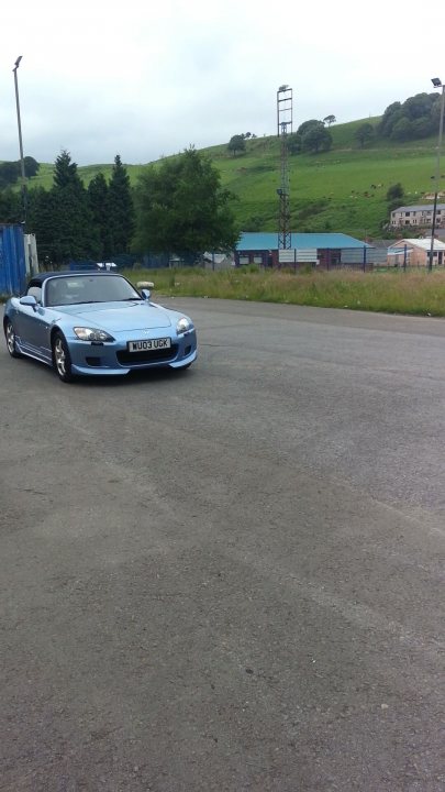 A car parked on the side of the road - Pistonheads - The image captures a scene of solitude. There is a blue sports car dominating the foreground. It's a sleek and shiny convertible, its top down, driving on a paved area that could be a street or a parking lot. 

Slightly blurred, suggesting a motion-effect, is a person on a bicycle, adding life to the otherwise serene setting. The background offers a contrast with elements of activity and nature - power lines stand against the gray sky, while lush green hills roll under the horizon. 

Additionally, there are traffic lights hinting at an upcoming controlled intersection, and a blue fabric structure, possibly an awning or fabric billboard, adds a dash of color to the scene. The overall image paints a picture of a peaceful ride amidst everyday human developments.