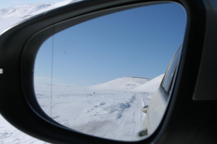 Iceland on a budget? - Page 1 - Holidays & Travel - PistonHeads - The image captures a breathtaking, ice-covered landscape through a side mirror of a car. The mirror reflected a long, snow-covered road leading up to a snowy hill. A gate can be seen in the distance along the road. The car's rearview mirror reflects the white expanse of snow around, creating a stark contrast between the heated interior and the cold exterior of the scene. The mirror frame, colored black, encapsulates this panoramic view, presenting it in a confined, square perspective. The entire scene exudes a sense of tranquility and isolation amidst the winter wonderland.