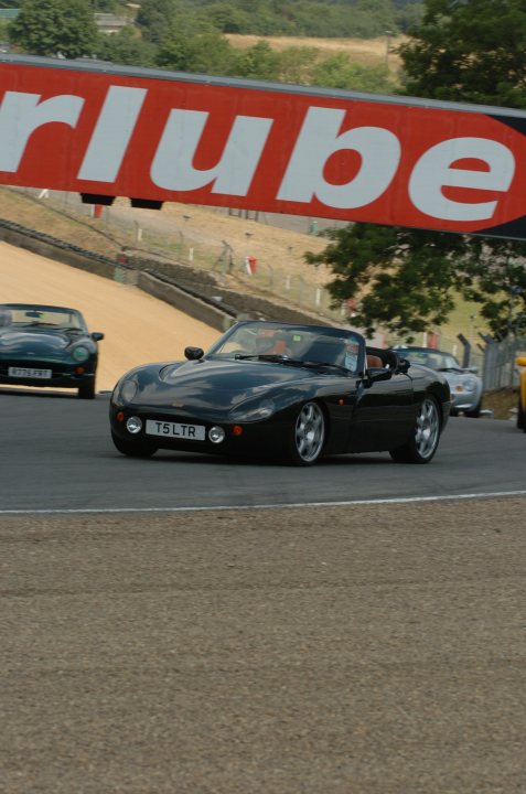 Random TVR pics - Page 36 - General TVR Stuff & Gossip - PistonHeads - This image depicts an exhilarating scene from a race track. The central focus is a sleek, black sports car with a European-style license plate, driving near the edge of the track. In the background, there's another car, possibly in the racing lineup, and a sizable banner displaying the word "Tonik" in bold letters. The track is set against a backdrop of lush green trees, enhancing the sense of speed and competition. The overall mood of the image is dynamic and competitive, showing the intensity and excitement of racing.