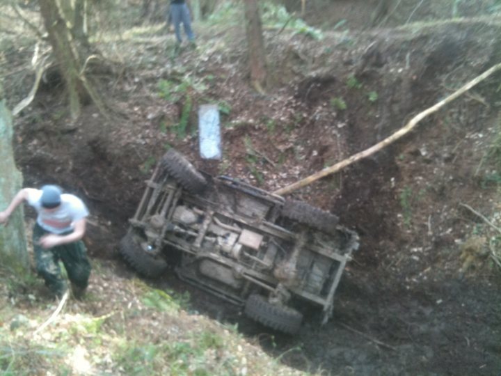 Pistonheads - The image captures a tense outdoor scene where a person in a purple shirt is leaning over, possibly inspecting, a ruined off-road vehicle that has tipped over onto its side. The vehicle is partially submerged in a muddy area, and the person seems focused on assessing or possibly fixing the situation. The vehicle's tires are prominently visible, indicating the challenging terrain. The environment around them is shady and covered in thick foliage, suggesting they could be in a wooded area or near a heavily vegetated place. The fallen tree trunk is likely a casualty of the vehicle's crash.