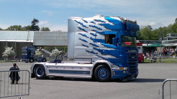 Long cabs in UK? - Page 1 - Commercial Break - PistonHeads - The image showcases a large blue truck, which is predominantly white with a fish design adorning its side. The truck appears to be quite prominent, with its large size and bold colors making it a focal point. It's parked on a black tarmac pavement, adding a stark contrast to the truck's colors. In the background, there are a variety of people and structures, including a building and a chair, suggesting this scene might be taking place in a parking or exhibition area. The lighting is bright, casting soft shadows and highlighting the truck's distinctive features.