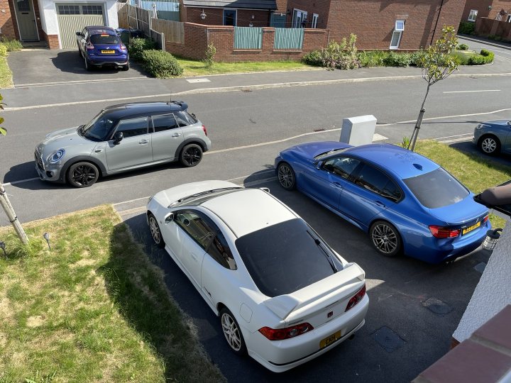 RE: Neverending two-car story | PH Footnote - Page 7 - General Gassing - PistonHeads UK - The image is a color photograph showing an outdoor scene of several cars parked in a driveway. There are various types and colors of vehicles, including sports cars and sedans. Some cars have visible sunroofs or convertible tops up. A few personal items like backpacks and handbags can be seen near the cars. The environment suggests it might be a residential area, as indicated by the presence of a house in the background and a driveway. There is also a hint of greenery at the edge of the frame. The sky appears overcast, and there are no people visible in the immediate vicinity of the cars.