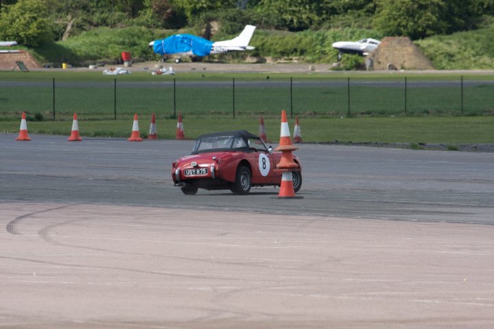 Spring London Summer North April Pistonheads - The image shows an orange pylon, presumably a traffic direction marker, situated in the center of the photograph. In the background, a vibrant red vehicle is seen, likely on a race track or a similar course given the presence of pylons. There's a fenced-in area to the left, and the scene appears to be outdoors during the day. A blue canopy is visible in the far distance, and the surrounding environment includes grassy areas and trees.