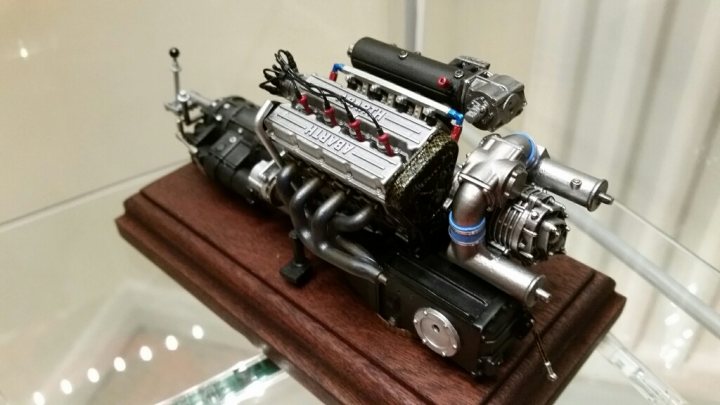 MFH 1/12 Lancia Delta S4 Engine - Page 1 - Scale Models - PistonHeads - The image shows a miniature model of a powerful V8 engine. The engine, a complex assembly of pipes and cylinders, dominates the scene with intricate details. The engine appears to be black and silver in color, mounted on a wooden base. On the engine model, there are labels showing the make and model, but they are not clearly legible. In the background, there is a wall with white tiles, creating a contrast with the engine on the brown display stand.