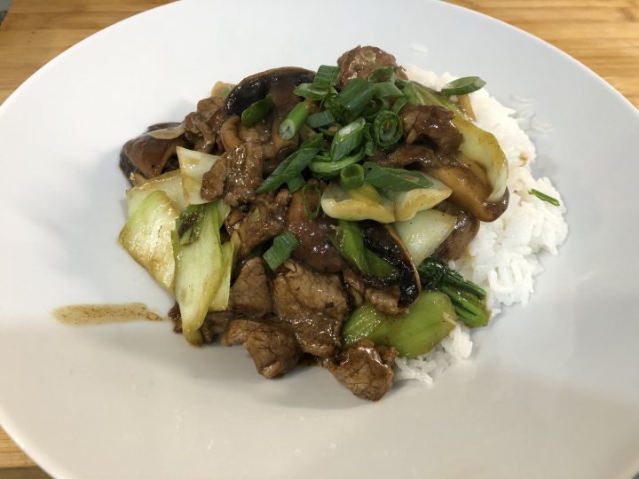 Pistonheads - The image shows a meal consisting of meat and vegetables served on a white plate. Accompanying the main dish is a serving of rice, possibly fried, garnished with chopped scallions. A sprig of what appears to be parsley is placed atop the food as decoration or flavor enhancer. The plate is set on a wooden surface, adding a rustic touch to the presentation. The meal looks like an Asian-style dish, possibly beef and broccoli, given its ingredients and arrangement.