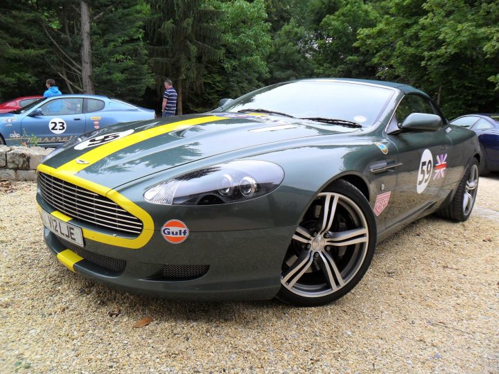 Aston Cars stickered up for LeMans. - Page 1 - Aston Martin - PistonHeads - The image shows a grey sports car parked on a gravel surface. The car is adorned with various design elements, such as yellow and red stripes, and it bears a large circular logo that says "GULF" and a smaller one at the back that likely reads "59." There are people in the background standing, but they are not the focus of the image. The setting appears to be an outdoor, daylight scene with a serene ambiance. The style of the image suggests it is a photograph.