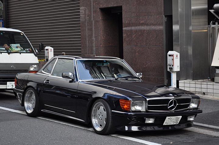 Pistonheads - The image depicts a black Mercedes-Benz sports car parked in a city street. The stop sign next to the car is blue and white. Another car can be seen parked behind the Mercedes, and there is a building with a reflective metallic panel in the background. The stop sign is placed next to the street where the car is parked. The Mercedes sports car has a distinctive license plate with the number 3 on it.