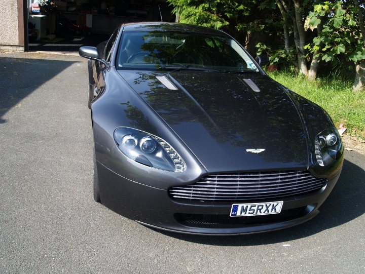 Please can we lighten up ? - Page 1 - Aston Martin - PistonHeads - The image shows a black Aston Martin luxury sports car parked on a pavement. The car's sleek design is prominent, with the hood and bodywork in a shiny black finish. The car has a distinctive rounded nose, iconic headlamps that taper towards the front, a grille with a fresh air intake, and angled side mirrors. The vehicle also has a license plate plate with the registration "MSSLXK," which is attached to the front bumper. The setting seems to be a residential area, as there is a house and trees in the background, contributing to a peaceful and exclusive ambiance.