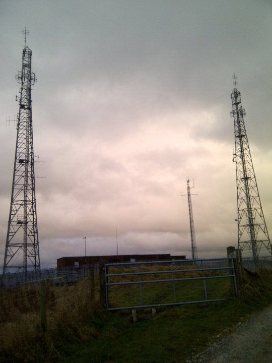 Anyone know anything about Tetra masts? - Page 1 - Science! - PistonHeads - The image features an open field with two tall metal towers and a building in the background. The towers and building are enclosed by a fence with posts sticking out of the ground. The sky is overcast, suggesting that the photo might have been taken on a cloudy or stormy day. The area is bordered by a dirt path leading into the distance, providing a sense of depth and scale to the scene.