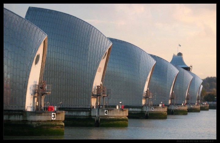 Pistonheads Piece Engineering Iconic Personalmost - The image showcases a unique arrangement of heavy, square metal structures resembling shields, lined up parallel to each other on the water's edge. They could possibly be enormous silos or part of an architectural installation. The protective walls of the structures are gleaming, and the reflections on the water's surface create a mirror-like effect, almost giving the illusion of doubled structures. The sky above is a soft, cloudy blue with a smattering of clouds clearing to reveal the sky.