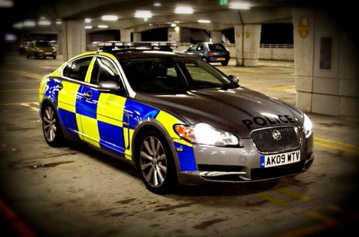 Buying Pistonheads British - The image captures a close-up view of the side of a silver Volvo police car, possibly a V50 model. The car is parked in a parking garage with a yellow-and-green checkered pattern on the hood. A "POLICE" sign is prominently displayed on the top of the car, alongside the registration number "AK09 TVP". The interior of the garage features tiered levels and poles, typical of such structures.