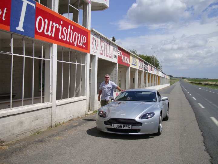 French trip - Page 1 - Aston Martin - PistonHeads - The image presents a vibrant scene from a storefront named "Touristique." The building stands out with its red and white paintwork. A man is seen standing outside the store, while a silver sports car is parked right in front on the street, which stretches out in the distance. The storefront features a colorful sign in the window above a display that advertises "Gloria Shoes." The architecture and wares suggest a nicely preserved establishment on a bustling street.