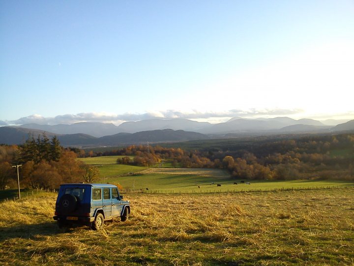 Best off road fun in Scotland - Page 1 - Off Road - PistonHeads - The image features a serene countryside scene. A blue vehicle, possibly a pickup or SUV, is parked at the lower portion of a lush green field. The vegetation appears to be a mix of grass and hay. Beyond the field, a forested valley or small village unfolds, leading the eye towards majestic, green-capped mountains in the distance. Above, the sky is a clear, bright blue, indicating either early morning or late afternoon, with a few white clouds scattered throughout. The overall scene conveys a sense of tranquility and natural beauty.