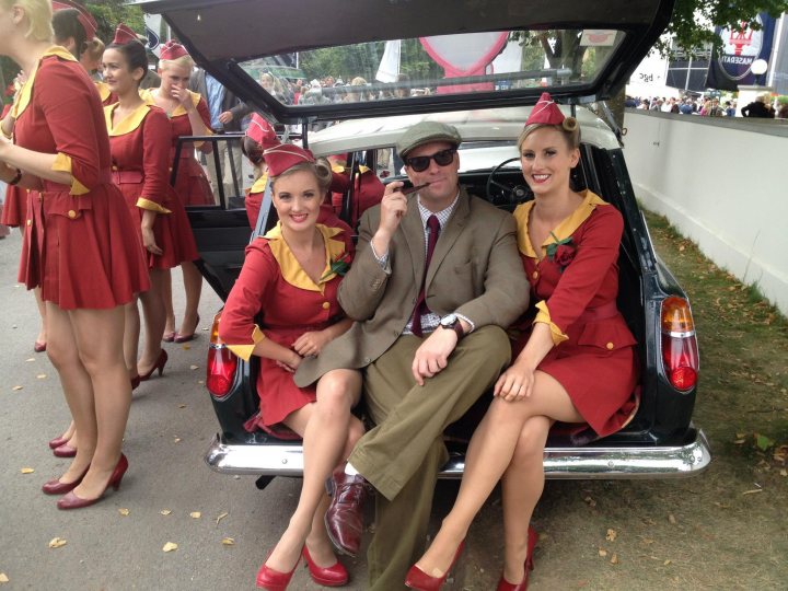 2012 Goodwood Revival Meeting Photo Gallery - Page 2 - Goodwood Events - PistonHeads - The image captures a scene from a car show or parade. In the heart of the action, a classic convertible car carries three chestnut-haired women in vibrant red dresses. They are seated comfortably, adding a touch of whimsy with their matching red heels. Adding to the charm, two of the women sport floppy red hats and all three have red bouquets, creating a harmonious visual. The man, presumably the driver, sits behind the steering wheel, blending the modern with the vintage by wearing a tie and glasses. The car appears to be the center of attention, with onlookers standing in close proximity, suggesting a sense of excitement and anticipation in the air.