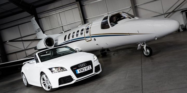 One photo of your current car - Page 1 - South West - PistonHeads - The image features a luxurious Audi R8 sports car and a large private jet set against a white corrugated metal wall. The jet, painted in white with blue stripes, occupies the background of the image. The Audi R8, with its white exterior and silver rims, is prominently positioned in the foreground. Both the jet and the car are parked in an industrial setting, suggesting a context of luxury and high-end travel.