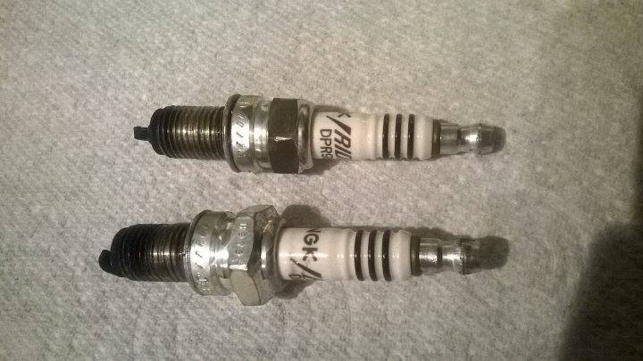 How remove incorrect spark plug i.e. no clearance - Page 1 - Home Mechanics - PistonHeads - The image is a close-up photograph of two cylindrical objects, which appear to be automotive spark plugs. These parts are metallic with gold-like casings, and one of them has lettering or numbers on it, which might indicate brand or model information. The plugs are positioned next to each other, and there is a texture that looks like a thread on both ends as they are not fully enclosed by a separate part like a hexagonal nut or a rubber gasket. The background is neutral and out of focus, emphasizing the subject matter. The photo captures their mechanical properties and design, typical for such automotive accessories.