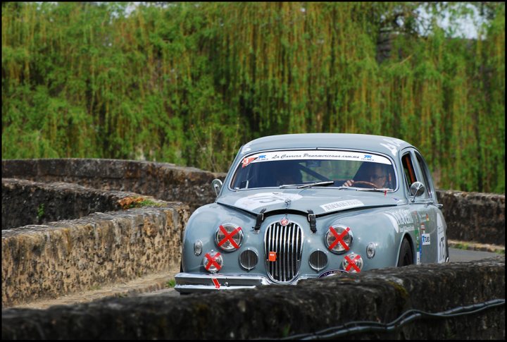 The Tour Auto came to town - Page 1 - Classic Cars and Yesterday's Heroes - PistonHeads - The image portrays a classic car, possibly a Citroen, racing on a curvy track. The car is predominantly gray, and it is marked with red crosses on the hood. The driver can be seen from behind the wheel, fully engaged in the drive. The car is driving on the right side of the road, and the track is bordered by lush green trees, creating a scenic backdrop for the vehicle.