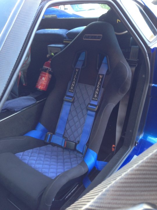 High Downforce Wing Risers - Page 1 - Noble - PistonHeads - The image captures the interior of a blue sportscar, showcasing a striking blue and black Sparco racing seat. The seat features shoulder belts and a red can, possibly containing fuel, adding to the sporty ambiance. Dominating the foreground is a close-up view of the seat's footwell, where a red fire extinguisher is prominently placed, hinting at a measure of safety equipment despite the intense racing setting.