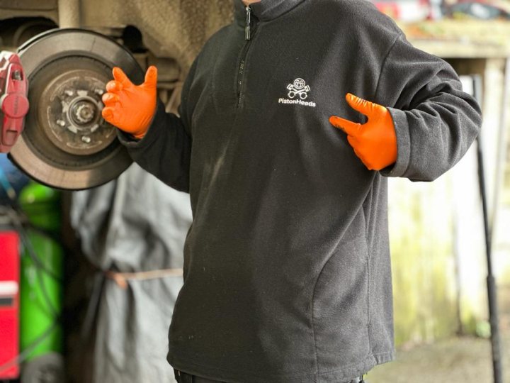 Pistonheads - The image captures a scene in a garage, where a person is engrossed in work. This individual, clad in protective gear, stands amidst various tools and equipment. Their arms are extended outwards, with one hand gesturing towards something unseen, while the other holds an orange object that appears to be a part of the mechanical process. The garage setting is characterized by several other individuals, each contributing to the bustling environment. Despite the focus on the central figure, the presence of others suggests a collaborative effort in this workspace.