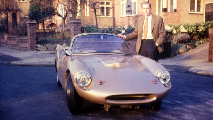 Ashley/Tornado Special.... - Page 1 - Classic Fibreglass - PistonHeads - The image features a man standing beside an old silver car with a convertible top. The man is dressed in a suit and tie, leaning on the vehicle. The car appears to be an older model, with a distinctive grille and round headlights. The setting is a residential street with houses on both sides, suggesting a suburban environment. The car's color and design suggest it might be from the first half of the 20th century.