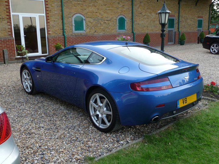 Finally took the plunge!  *happy bunny* - Page 1 - Aston Martin - PistonHeads - The image shows a blue sports car parked on a gravel driveway adjacent to a building with a brick facade and green window frames. The car features a two-door, coupe-style design with a sleek profile and a rear spoiler. A distinctive yellow license plate is visible. The driveway is well-maintained, and there are some potted plants and a lamp post along the pathway, which indicates a residential area. The sky appears to be clear, suggesting good weather conditions.