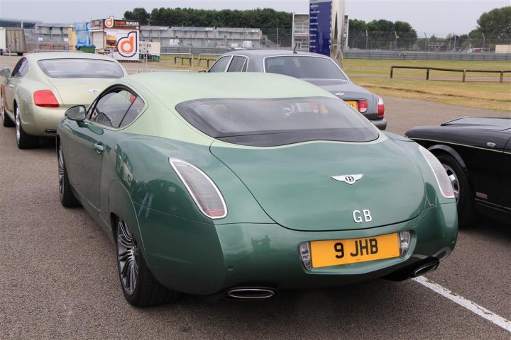 Strange Bentley - Page 1 - Rolls Royce & Bentley - PistonHeads - The image captures a scene at what appears to be a car show. The focal point is a striking green Aston Martin DB9, positioned towards the center. The car's sleek design and glossy finish suggest it is well-maintained. In the background, there are several other luxury cars on display, indicating the event's high-end nature. A stand with various items, likely merchandise or promotional material, is visible, adding to the event's ambiance. The overall setting seems to be on a paved road or tarmac, with a fence can be seen in the far background.