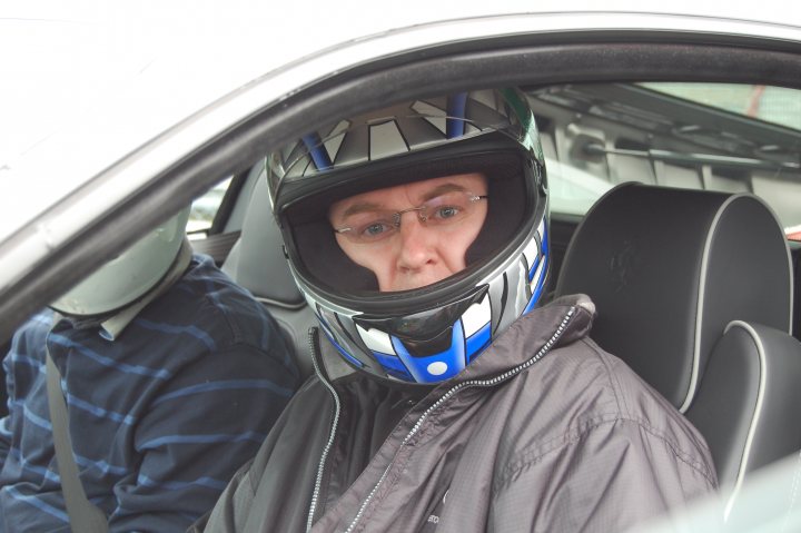 Castle Association Supercar Stroke Pistonheads Combe Day - The image features the interior of a vehicle where the camera is likely positioned near the door. In the foreground, a man is sitting in stark contrast to the black exterior. He is wearing a black and blue motorcycle helmet, suggesting he might be embarking on a ride. The man has a somewhat solemn expression on his face, possibly obscured by his helmet. Around him, the interior of the car is visible, including a black seatbelt and the car's upholstery. There is also a blue pattern on the man's shirt, adding a pop of color to his attire. His foot is resting on the car's pedal, further indicating the imminent motion of the vehicle. The image likely captures a moment just before a journey begins.