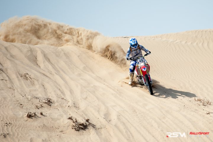 My Photos of the Year - Page 1 - Middle East - PistonHeads - The image captures an intense moment of a person riding a dirt bike across a sandy landscape, leaning forward to maintain balance as they navigate the terrain. The bike is kicked up with the rear wheel leaving a trail in the sand, indicating a sudden change in speed or direction. The rider is equipped in a white and blue motocross suit with protective gear, highlighting the adrenaline-fueled nature of the sport. The backdrop features a vast sand dune and a clear sky, adding a sense of adventure and freedom to the scene.