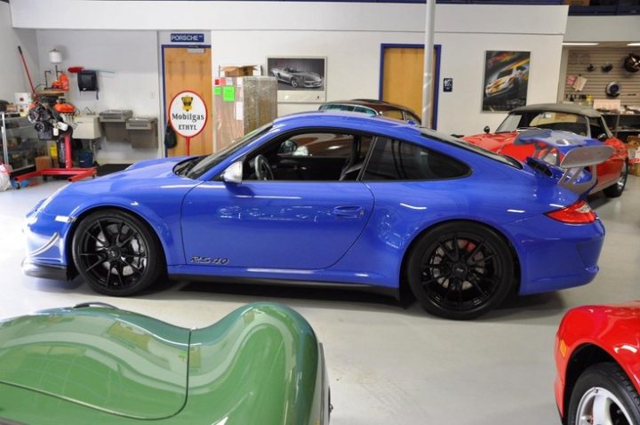 RS 4.0 values  - Page 12 - 911/Carrera GT - PistonHeads - The image shows a blue Porsche 911 GT2 racing car on display inside a car showroom. The car is sleek and polished with a unique paint scheme. It features the iconic Porsche design elements such as the low-slung body and large, theatrical rear wing. Positioned centrally in the photo, the car is the main focal point. Surrounding it are various other cars, hinting at the diverse selection of vehicles at the showroom. The lighting is bright, enhancing the vibrant color and shiny surfaces of the cars.