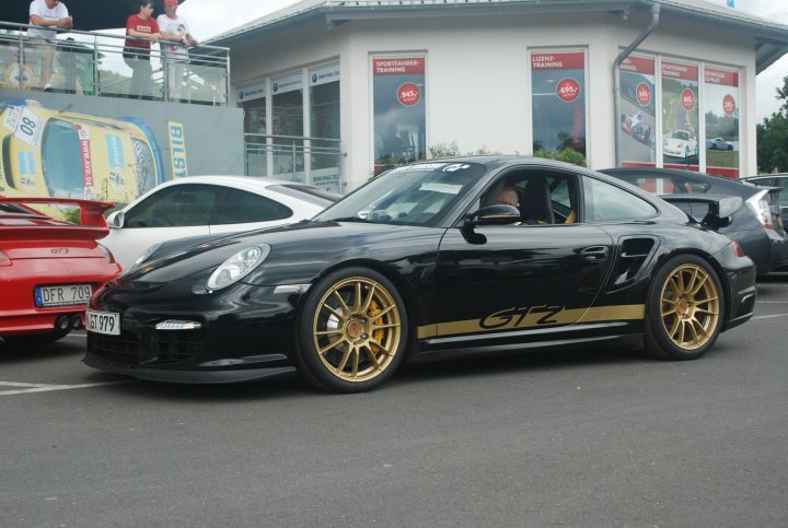 A car is parked on the side of the road - Pistonheads - The image shows a black Porsche sports car parked in front of a white building with tinted windows and a red awning. The car has distinctive 18-inch gold alloy wheels and the Porsche logo is visible on the hood. Behind the sports car, there is a line of four other vehicles parked orderly next to each other, waiting at a parking lot restroom. In the background, there are white trucks and a red car, suggesting a commercial or public parking area.