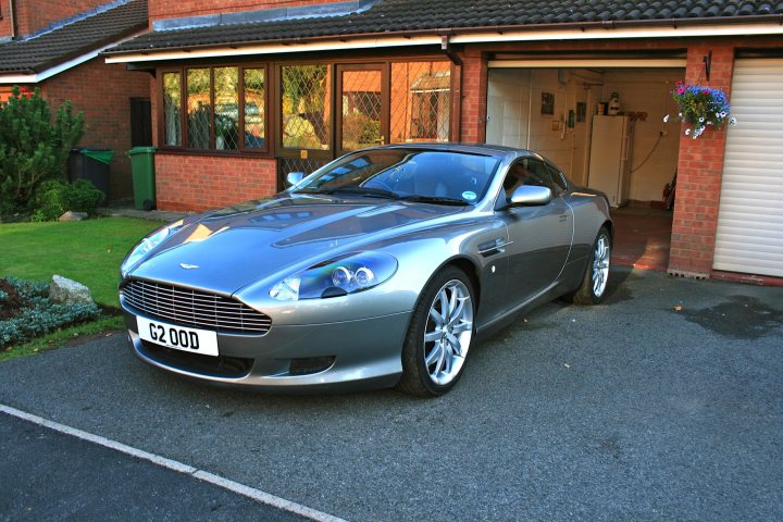 My First Aston - Page 1 - Aston Martin - PistonHeads - This image depicts a sleek, silver-colored Aston Martin car parked on a driveway in front of a brick house with a double garage. The car features a paddle gear shift, a vertical grille, and silver alloy wheels. There is a license plate visible on the car, indicating a UK registration. The house has a well-maintained garden area with bright green grass and a narrow path leading to a protected porch area. A purple hanging basket can be seen on the right side of the entrance. The driveway seems well-kept, and there is a brick wall on the left side of the frame, likely belonging to the neighboring house.