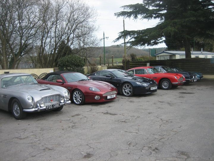 Owners Pistonheads - The image features a row of classic cars parked on a concrete surface. Starting from the left, there is a light blue car, followed by a striking red car, then a sleek black car, and finally a shiny silver car on the far right. The cars are aligned facing the same direction, suggesting attendees at an event or a car show. The setting appears to be outdoors, possibly in a parking lot, based on the no-frills background that includes a sign and skeletal trees.