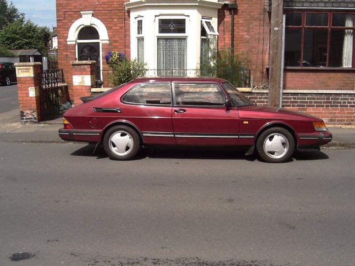 Saab 900 from 1992 suspension freshup - Page 1 - Saab - PistonHeads - In the image, a vintage red sedan is parked on the side of a street in a residential area. The car, which is a classic or newer model, is stationed in front of a house that has a charming white cottage aesthetic. The side of the street where the car is parked is flanked by brick houses, adding to the residential feel of the scene. There are no people or moving vehicles visible in the image, giving it a calm and still atmosphere.