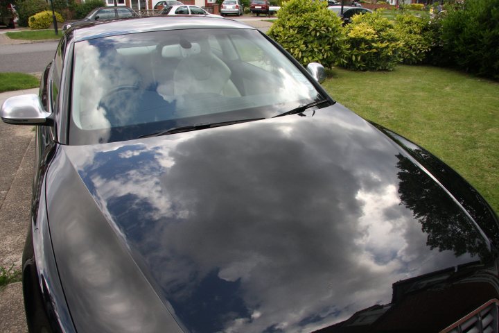 Audi S5 (pictures) - Page 1 - Audi, VW, Seat & Skoda - PistonHeads - The image captures a dark-colored Prospect Park car parked on a sunny day. The car's windshield offers a reflection of its surroundings. In the background, neatly trimmed bushes and a solitary tree provide a serene suburban backdrop, while other cars can be seen parked further back. The sky is filled with fluffy clouds, casting a soft, diffuse light that illuminates the scene.