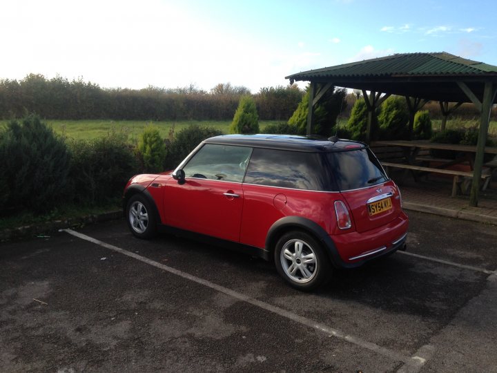 £3k - lease or buy, looking for a deal. - Page 1 - General Gassing - PistonHeads - The image shows a red Mini Cooper parked in a parking lot. The car is facing towards the right side of the image, parked next to a green pole. The parking lot appears to be near a grassy area, and there's a covered picnic area in the background, suggesting the location might be a park or a public garden. The overall scene is calm with no visible people or other moving objects. The car's license plate is clearly visible.