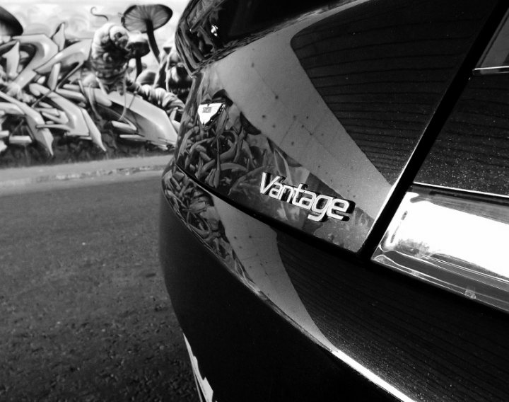 A black and white photo of a parking meter - Pistonheads - This image is a close-up of the door of a vehicle, focusing on the name "Vantage" embossed on the door. The text is clear, with a reflection visible in the glasses of the car. In the background, there's a black and white mural of people and trees, giving a contrast to the vehicle's sleek finish. The overall style corresponds to a dynamic urban setting.