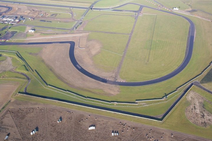 Pistonheads Snetterton - The image is an aerial photograph of a large, curved racing track situated in an open, flat landscape. There are several smaller vehicles near the track, likely part of maintenance or preparation activities. Grassy fields enclose the track, suggesting the complexity of the course with different lanes. The infrastructure hints at the scale of the raceway, designed for vehicular racing.