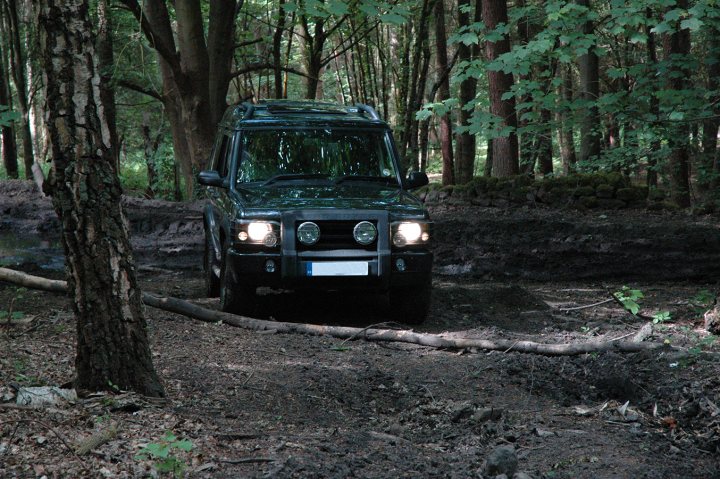 RE: Porsche Cayenne S: Spotted - Page 2 - General Gassing - PistonHeads - The image shows a rugged black SUV parked in a forested area. The vehicle is situated near the woods, with trees encroaching on its surroundings. There is no clear indication of a water source visible in the scene, but the terrain appears to be compacted and muddy, possibly where a log has been placed on the ground. A different pathway seems to be hedging through the treeline in the background, offering a potentially alternative route. The lighting suggests this could be early morning or late afternoon.