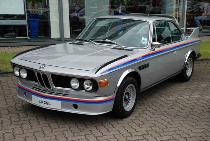 Sunday Pistonheads Service - The image shows a vintage car set against a modern city backdrop. The car, which is prominently displayed, is a gray BMW, identifiable by the blue and red stripes on its aerodynamic hood. It has a sleek, two-door body and features Bavarian automobile manufacturer BMW's distinctive emblem on the hood. The car is parked on a cobblestone street, suggesting an urban setting, and there are spectators and other cars in the background, indicating that this might be a city street or an event.