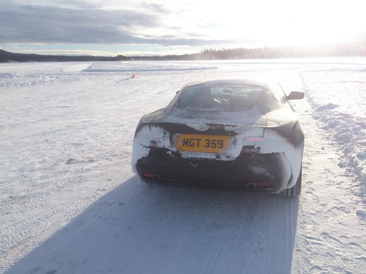 Aston Martin DB9 on Ice lake - Page 1 - Aston Martin - PistonHeads - The image showcases a serene winter scene. A car, with a yellow license plate and the registration number "GT359," is parked on a snow-covered road. The car is behind and to the right of a road marker. The road appears to be near an open track, which suggests the car might have been used there, given its mudflaps. The sky overhead is overcast, casting a soft light over the scene. The overall impression is of a quiet, possibly isolated, location perhaps after a period of vehicle activity.